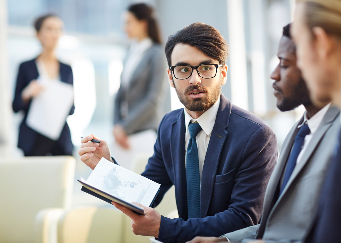 Three men in suits in the foreground discuss sales strategy. Two business women stand and talk in the background.